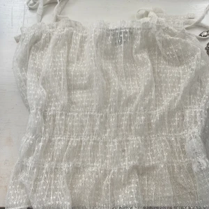 Vit spetsig linnetopp från Primark - Supersöt vit linnetopp från Primark med tunna axelband och transparent spetsmaterial. Toppen har smockad midja och ett diskret prickigt mönster i tyget. Perfekt för en somrig look med romantiska vibes.