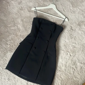 Svart axelbandslös kort klänning - Snygg svart axelbandslös kort klänning med dubbelknäppning framtill och en väldigt elegant passform . 