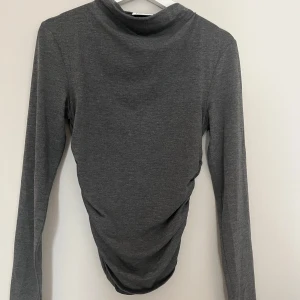 Grå långärmad topp från Kappahl - Säljer en grå långärmad topp från Kappahl i mjuk bomullsmix. Toppen har en snygg rynkad detalj i sidan som ger en cool twist och en lite mer figurnära passform. Perfekt att styla med jeans eller kjol för en chill look.