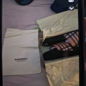 Burberry sandaler med klassiskt rutmönster - Säljer ett par snygga sandaler från Burberry med det ikoniska beige, svarta och röda rutmönstret. Remmen är i syntetmaterial och sulan är svart. Perfekta för dig som vill ha en lyxig och trendig look till sommaren.