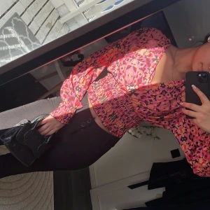 Färgglad blus med mönster - Säljer en färgglad blus med ett livligt mönster i rosa, rött och orange. Blusen har långa ärmar och en smockad midja som ger en fin passform. 