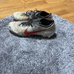 Nike fotbollsskor med röd swoosh - Snygga fotbollsskor från Nike med en iögonfallande röd swoosh på sidan. Skorna har en grå och svart färgkombination med snörning för optimal passform. Perfekta för fotbollsplanen!