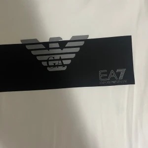 Vit t-shirt från Emporio Armani - Snygg vit t-shirt från Emporio Armani med ett svart band över bröstet och det ikoniska EA7-logotypen. Perfekt för en stilren och modern look. Kortärmad och tillverkad i mjukt material för extra komfort.