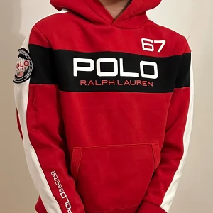 Röd hoodie från Ralph Lauren - Snygg röd hoodie från Ralph Lauren med svart och vit detaljering. Den har en stor POLO-logga på bröstet och nummer 67 tryckt på framsidan. Ärmarna har vita inslag och en POLO Racing-märke på sidan. Perfekt för en sportig look!