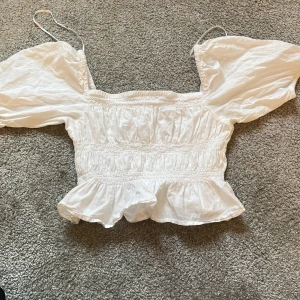 Vit blus från Gina Tricot - Säljer en söt vit blus från Gina Tricot med puffiga kortärmar och smockdetaljer. Blusen har en volangkant nedtill. Liten miss färgning under armen men syns inte så mycket