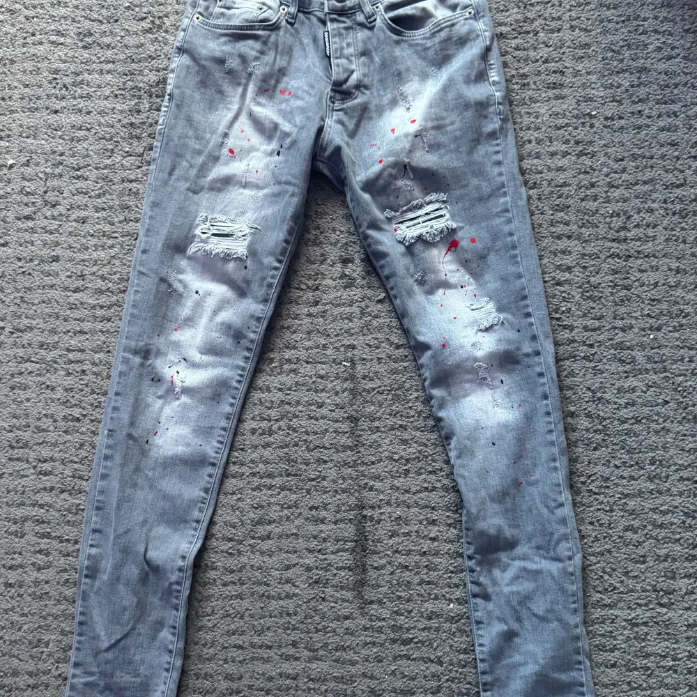 Snygga grå jeans från Dsquared2 med unika färgstänk i rött och svart. Byxorna har en slim passform och är dekorerade med slitningar på framsidan. De har en klassisk femficksdesign och knappgylf. Perfekta för en trendig look!. Farkut & Housut.