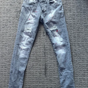 Dsquared2 jeans - Snygga grå jeans från Dsquared2 med unika färgstänk i rött och svart. Byxorna har en slim passform och är dekorerade med slitningar på framsidan. De har en klassisk femficksdesign och knappgylf. Perfekta för en trendig look!
