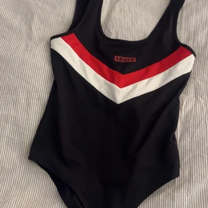 Svart body från Levi's - Snygg svart baddräkt från Levi's med ett coolt rött och vitt V-format mönster framtill. Perfekt för en dag på stranden eller vid poolen. Tillverkad i ett bekvämt material som ger en bra passform.
