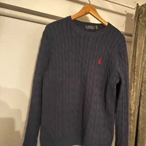 Mörkblå stickad tröja från Polo Ralph Lauren - Säljer en stilren mörkblå stickad tröja från Polo Ralph Lauren med kabelstickat mönster. Tröjan har en liten röd logotyp på bröstet och långa ärmar. Perfekt för kyligare dagar!