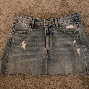 Jeanskjol med slitningar - Snygg jeanskjol i blå denim med slitningar och rå kant. Kjolen har en klassisk femficksdesign och stängs med knapp och dragkedja framtill. Perfekt för en avslappnad stil.