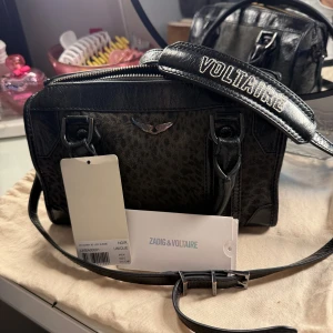 Svart handväska från Zadig & Voltaire - Snygg svart handväska från Zadig & Voltaire med leopardmönster och coola detaljer. Väskan har en justerbar axelrem med text och dragkedja upptill. Väskan är köpt i Portugal men säljer då jag inte längre använder den. Nypris: 4899 kr, mitt pris: 2000