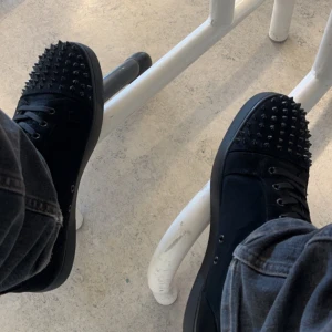 Svarta Christian louboutin sneakers med nitar - Snygga svarta sneakers med nitar på ovansidan och en ikonisk röd sula. Skorna har snörning och är tillverkade i mocka, vilket ger en lyxig känsla. Perfekta för att ge din outfit en edgy touch.