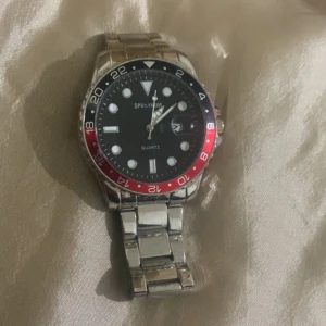 Klocka från Spectrum med stållänk - Snygg klocka från Spectrum med en stilig stållänk och en boett i rostfritt stål. Urtavlan är svart med tydliga markeringar och datumvisning. Bezel i rött och blått ger en sportig touch. Perfekt för dig som gillar en klassisk men ändå modern stil.