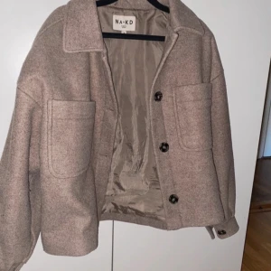 Beige jacka från NA-KD - Säljer en stilren beige jacka från NA-KD med stora fickor och knappar framtill. Jackan är perfekt för höst och vår. 