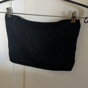 Svart glittrig tubtopp - Snygg svart tubtopp med glittrande detaljer. Perfekt för en kväll ute eller festliga tillfällen. Enkel och stilren design som passar till många olika outfits.