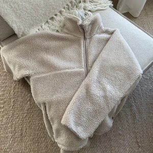 Beige teddyjacka med dragkedja - Mysig beige teddyjacka med hög krage och dragkedja framtill. Perfekt för kyliga dagar med sin fluffiga och varma design. Jackan har långa ärmar och en avslappnad passform.