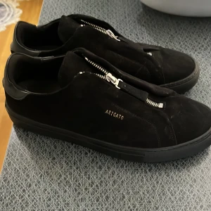 Svarta sneakers från Axel Arigato - Snygga svarta sneakers från Axel Arigato i mocka med dragkedja framtill. Perfekta för en stilren look. Diskret logga i guld på sidan ger en exklusiv touch.