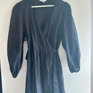 Svart omlottklänning från H&M - Snygg svart omlottklänning från H&M i linneblandning. Klänningen har långa ärmar och en v-ringad design som ger en elegant look. Perfekt för olika tillfällen och lätt att matcha med accessoarer.