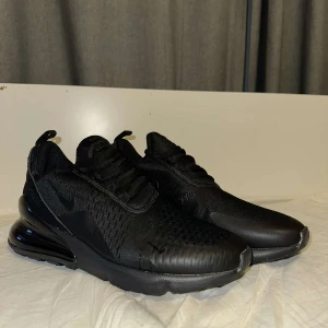 Svarta Nike Air Max 270 - Snygga svarta Nike Air Max sneakers med en stilren design. Skorna har en luftig ovandel i syntetmaterial och en synlig Air Max-dämpning i hälen för extra komfort. Perfekta för både träning och vardag.