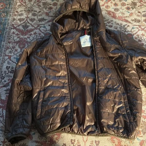  dunjacka från Moncler - Säljer en brun dunjacka från Moncler med huva och dragkedja. Jackan har ett quiltat mönster och är perfekt för kyligare väder. Den har en cool serietidningsdetalj på insidan. Perfekt för dig som vill ha stil och funktion i ett!