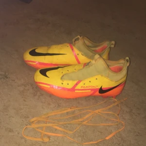 Gula Nike Phantom fotbollsskor  - Säljer ett par gula Nike Phantom fotbollsskor men oranga och svarta detaljer. Skorna har en svart Nike-logga och är designade för optimal prestanda på planen. Skorna har en modern design med snöring och är perfekta för fotbollsplanen. De har en slitstark sula med dobbar för bra grepp. Fotbollsskorna är storlek 36 men rekommenderar att inte köpa om man har exakt 36 då de är små i passform. Perfekta för dig som vill sticka ut!