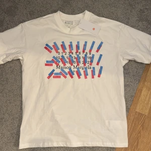Vit t-shirt från Maison Margiela - Säljer en tvär fet vit t-shirt från Maison Margiela med ett unikt tryck i blått och rött. Storleken är M men skulle säga att den passar mer som L. Den är i 10/10 skick (helt ny). Säljer för att den var för stor. Skriv vid funderingar!💯