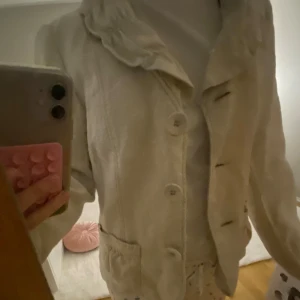 Beige kavaj med volangdetaljer - Säljer en stilren beige kavaj med volangdetaljer vid kragen och knappar framtill. Perfekt för en elegant look. Kavajen har långa ärmar och två fickor framtill. Köpt på loppis, men oanvänd av mig. Dock tog jag ur lite tyg från axlarna och därför är det synligare sömmar där men inget som syns mycket☺️☺️☺️
