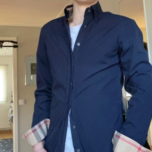 Mörkblå skjorta från Burberry Brit - Snygg mörkblå skjorta från Burberry Brit med klassiskt rutigt mönster på insidan av kragen och ärmsluten. Skjortan har långa ärmar och knappar framtill. Perfekt för en stilren look.