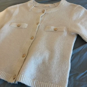 Vit kofta från Gina Tricot - Säljer en stilren vit kofta från Gina Tricot med korta ärmar och dekorativa guldknappar. Koftan har två fickdetaljer framtill och ribbade kanter för en snygg finish. Perfekt för en chic och avslappnad look.