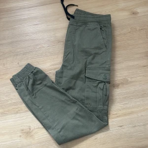 Gröna cargopants med snörning - Snygga gröna cargopants med elastisk midja och snörning. Byxorna har flera praktiska fickor med knappar och elastiska muddar vid bensluten. Perfekta för en avslappnad stil.
