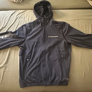 Mörkblå vindjacka från Lacoste - Säljer en stilren mörkblå vindjacka från Lacoste med dragkedja och huva. Jackan har långa ärmar och diskreta logotyper på bröstet och ärmen. Perfekt för blåsiga dagar och ger en sportig look., helt ny med prislapp, går att mötas eller fraktas!.