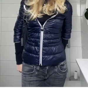 Mörkblå pufferjacka med dragkedja - Snygg mörkblå pufferjacka med dragkedja framtill. Jackan har en figurnära passform och är trekvartsärmad. säljer pga av att den ej kommer till användning. (lånade bilder från tidigare köpare på plick) 
