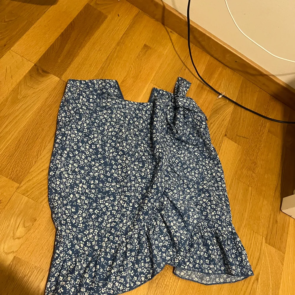 Säljer en söt blå kortkjol med vita blommor och volangdetaljer. Kjolen har en omlott-design med knytning i midjan för en justerbar passform. Perfekt för sommarens alla tillfällen!. Hameet.