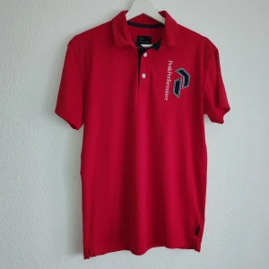 Röd pikétröja från Peak Performance - Snygg röd pikétröja från Peak Performance med broderad logga på bröstet. Tröjan har en klassisk krage och knappar framtill. Perfekt för en sportig look eller en avslappnad dag.