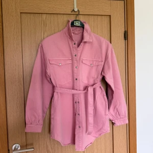 Rosa overshirt med löst bälte från Vila - Snygg rosa overshirt från Vila i bomull med bälte i midjan. Skjortan har långa ärmar, tryckknappar framtill och två bröstfickor med lock. Perfekt att styla lager på lager för en trendig look.