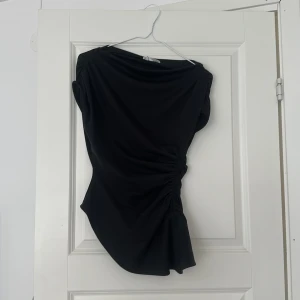 Svart asymmetrisk topp  - Svart topp från Zara med asymmetrisk skärning och snygg rynkdetalj på sidan. Modellen är ärmlös Tillverkad i mjukt, stretchigt material som sitter skönt på kroppen. Helt ny, lappen sitter till och med kvar, skulle lämna tillbaka då jag tyckte den var för stor men glömde bort att skicka tillbaka i tid. 