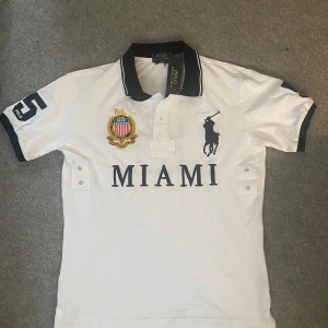Miami Polo Ralph Lauren Vit pikétröja - Vit pikétröja från Polo Ralph Lauren med mörkblå detaljer, broderad logga och Miami-tryck framtill. Klassisk krage med knappar, broderat emblem på bröstet och stor siffra 5 på ärmen och ryggen. 