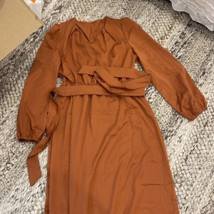 Rostbrun/orange långklänning med bälte - Snygg rostbrun långklänning med v-ringning och långa ärmar. Klänningen har ett matchande bälte i midjan. Storlek S. Satin
