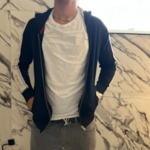 Svart hoodiejacka från GANT med logga - Svart hoodiejacka från GANT med vita ränder och GANT-logga längs ärmarna. Jackan har dragkedja framtill, huva och ribbade muddar. Perfekt för en chill och sportig stil. Materialet är mjukt och bekvämt, troligen bomull eller polyester.