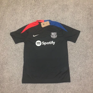FC Barcelona svart Nike fotbollströja - Svart FC Barcelona fotbollströja från Nike med vita loggor och sponsortryck. Tröjan har korta ärmar och detaljer i rött och blått på axlarna. Materialet är lätt och andas, perfekt för träning eller match. Matchande svarta shorts med klubbmärke och färgdetaljer ingår.