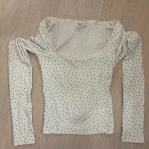 Vit blommig långärmad topp H&M XS - Supersöt långärmad topp från H&M Basic i vitt med små blå blommor. Toppen har en bred, rundad urringning med spetskant och är tillverkad i ribbad bomull. Perfekt för dig som gillar romantiska och feminina detaljer.