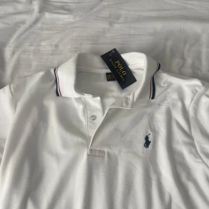 Vit pikétröja från Polo Ralph Lauren - Vit pikétröja från Polo Ralph Lauren med klassisk krage och knäppning framtill. Tröjan har marinblå logga broderad på bröstet och tunna ränder i rött och marinblått på kragen. Tillverkad i mjuk bomull, perfekt för en clean och sportig look.