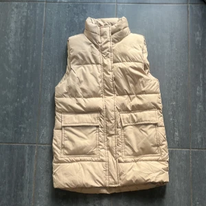 Beige dunväst från 157 - Snygg beige dunväst från 157 med hög krage och två stora fickor framtill. Västen är quiltad och har en stilren look som passar perfekt till lager-på-lager. 