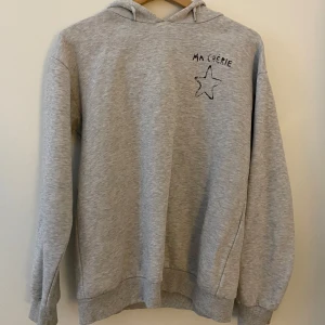 Grå hoodie från Gina Tricot  - Grå hoodie från Gina Tricot med broderad svart stjärna och texten 'MA CHERIE' på bröstet. Hoodien är mjuk och skön. Den är använd och tvättad ett flertal gånger men är fortfarande i bra skick💛💛