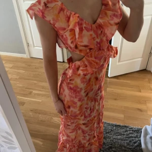 Färgglad långklänning med volang - Säljer en superfin långklänning i orange, rosa och gula toner med blommigt mönster. Klänningen har volangdetaljer och öppen rygg. Den är eftertraktad och slutsåld överallt. Så fin till sommaren. Pris går att diskutera vid snabb affär