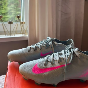 Nike fotbollsskor ljusgrå/rosa - Nästintill oanvända fotbollsskor. Nike Mercurial Vapor 16 Pro, strl 39 Säljer ett par fotbollsskor i toppskick , använda endast en gång på en träning innan min syster bestämde sig för att sluta.  • Modell: Nike Air Zoom Mercurial Vapor 16 Pro • Färg: Ocean Cube/Pink Blast •  Storlek: 39 • Underlag: Firm ground (FG – fasta gräsplaner)  Skorna är i princip som nya. Inga fläckar, skador eller slitage – endast lite märken från dubbarna som uppstår direkt vid användning. Nypris: 1999 kr