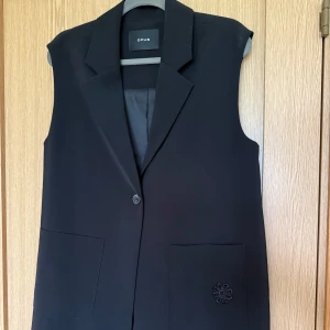 Svart lång väst från Opus - Stilren svart väst från Opus med klassisk krage och en knapp framtill. Västen är lång, har två stora fickor fram och en diskret broderad blomdetalj på ena fickan. Perfekt för lager-på-lager och ger en clean look.