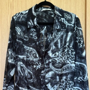 Svart paisleymönstrad skjorta Dranella - Snygg svart och grå blus från Dranella med paisleymönster och transparent känsla. Skjortan har långa ärmar, klassisk krage och råa metallknappar framtill. Materialet är lätt och luftig polyester med insydda rektangulära detaljer och guldfärgad tråd som ger extra struktur och lyx. 