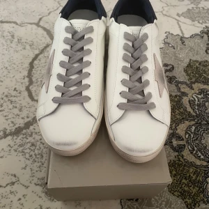 Golden Goose vita sneakers med stjärna - ETT AV ETT Golden Goose sneakers i vitt skinn med grå snörning och ikonisk grå stjärna på sidan. Baksidan har mörkblå hälkappa och detaljer i beige mocka. Snygg, avslappnad look med platt sula och rund tå. Perfekta för dig som gillar streetstyle och premiumkänsla. 