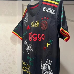 Ajax x Bob Marley Adidas fotbollströja - Snygg svart Ajax fotbollströja från Adidas med Bob Marley-tema. Flera färgglada tryck, Ajax-logga, Ziggo sponsor, och citat. Röd, gul och grön detalj på ärmar och axlar. Kortärmad och tillverkad i lätt, ventilerande material. Perfekt för dig som gillar unik stil och fotboll.